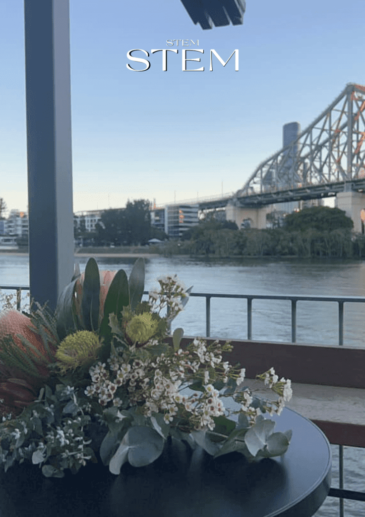 Native floral arrangement with proteas, leucadendrons, and wax flowers on a black table, overlooking the Brisbane River and Story Bridge at Howard Smith Wharves.