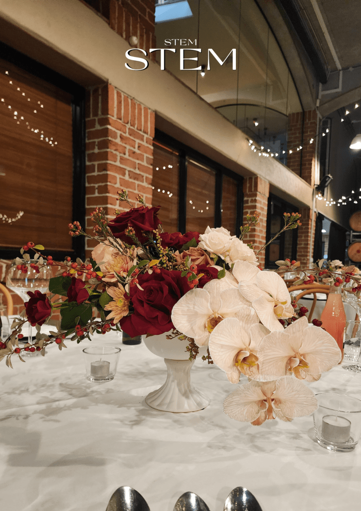 Bohemian chic wedding flowers by Stem Design featuring burgundy roses, blush orchids, and textured foliage in a white vase at a Brisbane wedding.