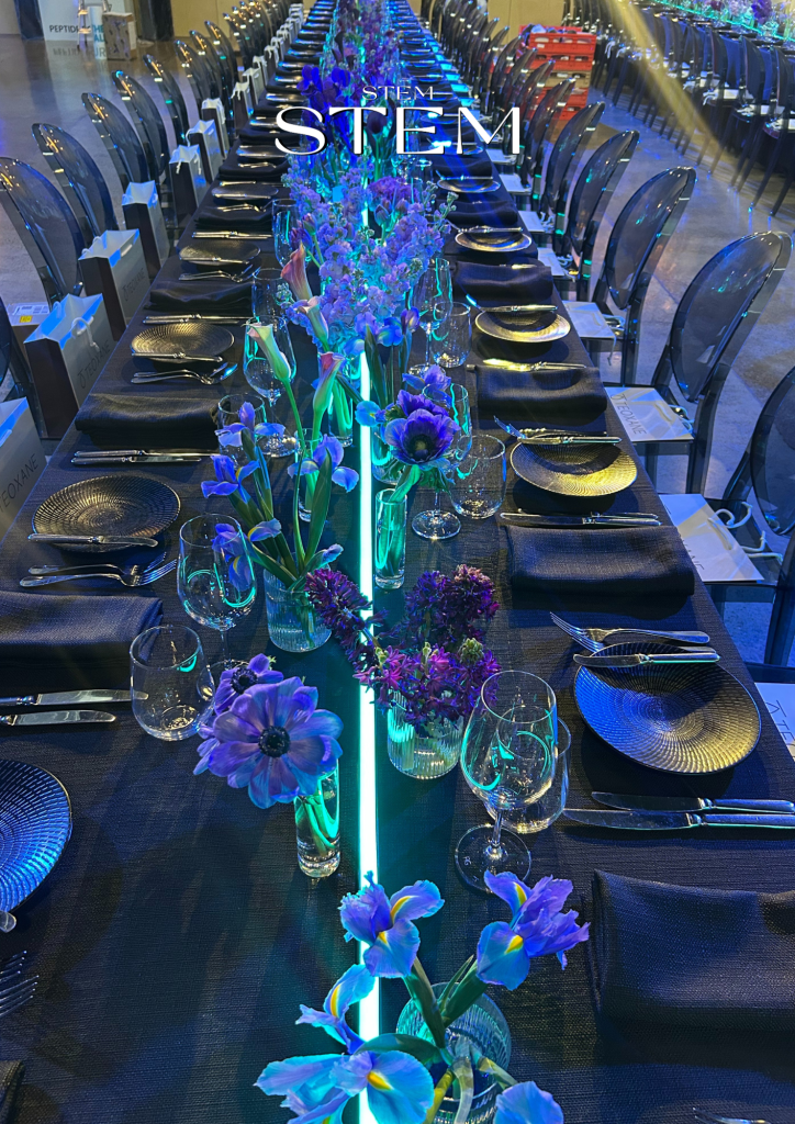 A long banquet table styled by Stem Design with glowing vases filled with purple and blue flowers, creating a dramatic illuminated effect for a Gold Coast event.